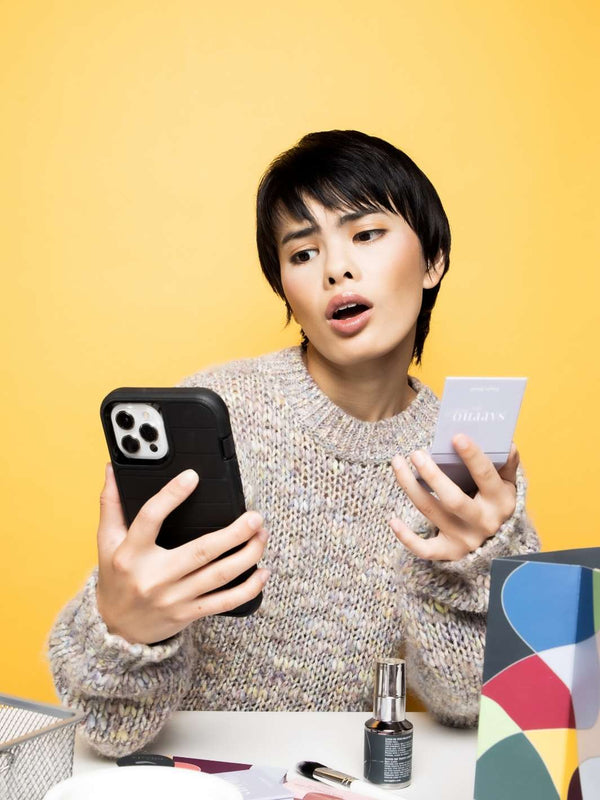 Asian woman having a video call on cell phone talking while holding a refillable compact during a virtual consultation with makeup artist sitting at desk with organic and sustainable beauty products scattered on her desk