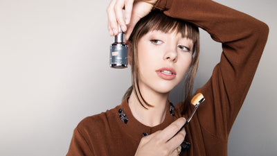 Model glancing sideways, looking at organic foundation bottle in her hand, cruelty free buffer brush in the other hand and wearing a rusty brown sweater with a natural makeup look and trendy fall 2021 hairstyle with bangs